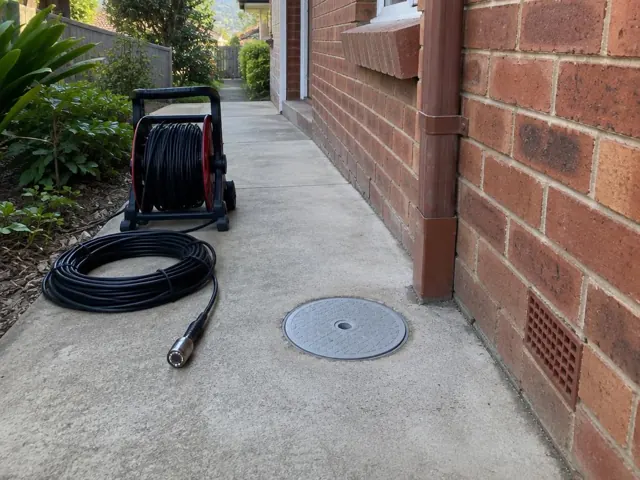 Capped drain inspection opening beside a coiled camera reel on a concrete side path by a brick wall in Helensburgh